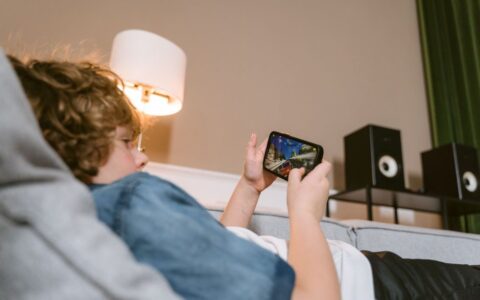 Teen lying on sofa playing video games on smartphone indoors, surrounded by modern décor.