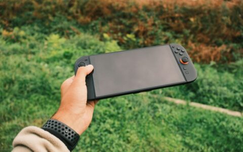 Close-up of a person's hand holding a black handheld gaming console against a green outdoor background.