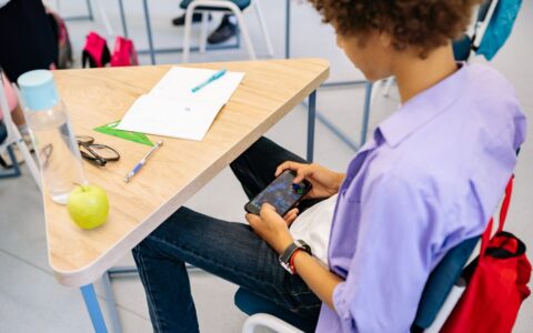 A child playing a mobile game at a school desk with school supplies nearby, captured indoors during class.