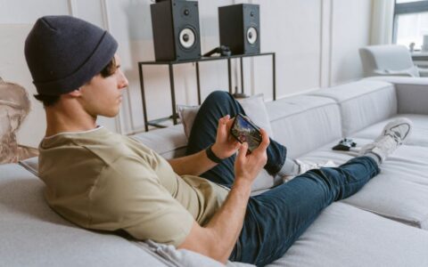 Casual young man playing mobile games on sofa with speakers in the background.