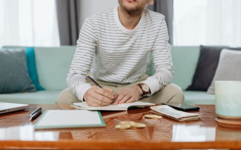 A man sits at a table writing in a notebook, managing finances and budgeting.
