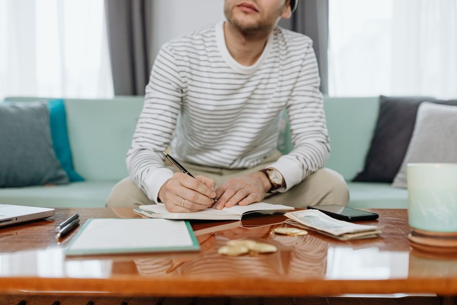 A man sits at a table writing in a notebook, managing finances and budgeting.