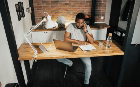 A young man at a desk in a home office, using a laptop and phone, embodying remote work lifestyle.