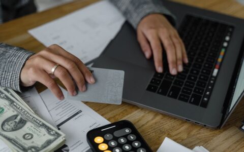 Close-up of a person using a credit card and laptop in a financial setting with cash and calculator.