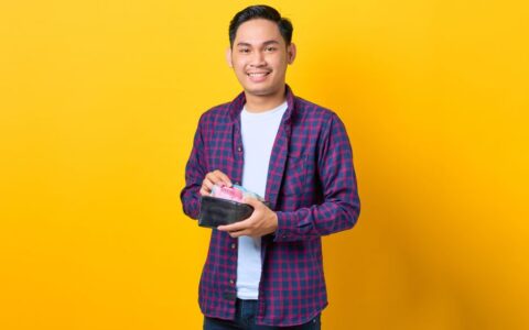 Happy young man in plaid shirt holding wallet with Indonesian rupiah against yellow background.