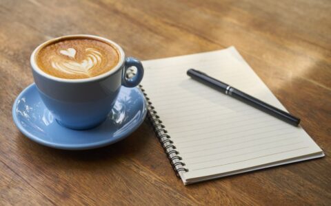 A calming scene of a latte with art in a blue cup beside a notebook and pen on a wooden table.