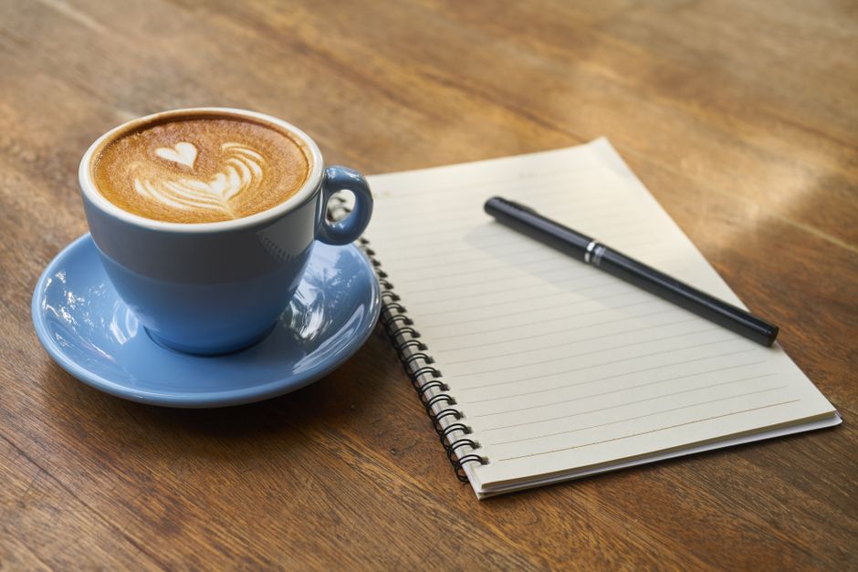 A calming scene of a latte with art in a blue cup beside a notebook and pen on a wooden table.