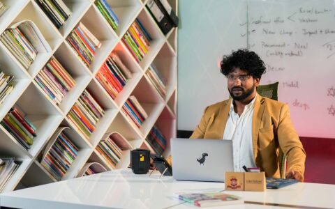Professional man in a modern office setting sitting at a desk with bookshelves and a laptop, appearing focused.
