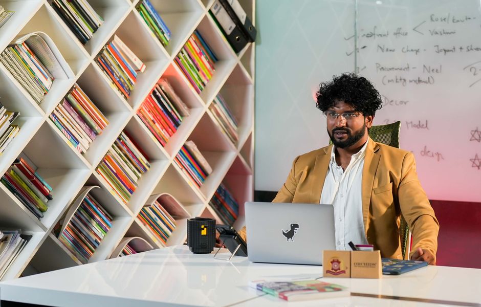 Professional man in a modern office setting sitting at a desk with bookshelves and a laptop, appearing focused.